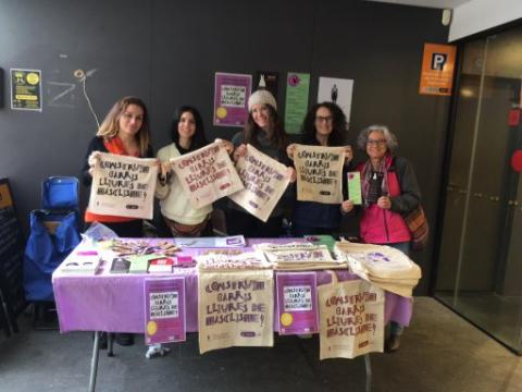 Fotografia de 5 dones que sostenen una bossa amb el lema "Construïm barris lliures de masclisme", darrere d'una taula amb diferents materials de difusió