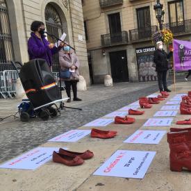 Fotografi on apareix en primera plana 19 parells de sabates vermelles amb els noms de les dones assassinades el 2021 i al fons, una dona parlant al micròfon.